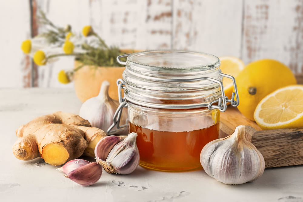 Glass jar with honey, ginger, lemon, garlic and camomile over bright grey background