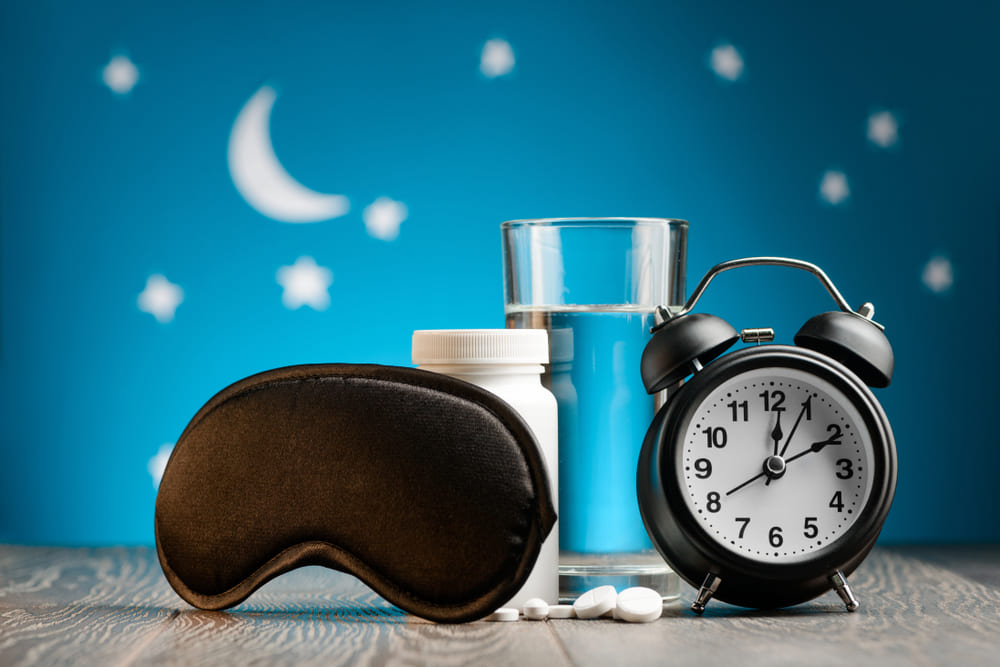 Eye mask, pills and alarm clock on a bedside table over night sky.