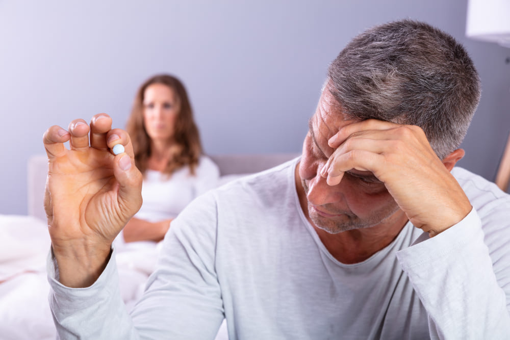 Matured Man Suffering From Erectile Dysfunction Holding Pill In Hand And His Wife In Background